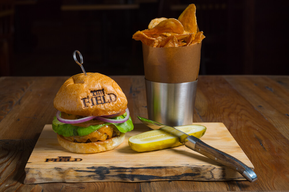 A burgerwith cheese, lettuce & onlin on a branded bun; homemade potato chips and a pickle spear.